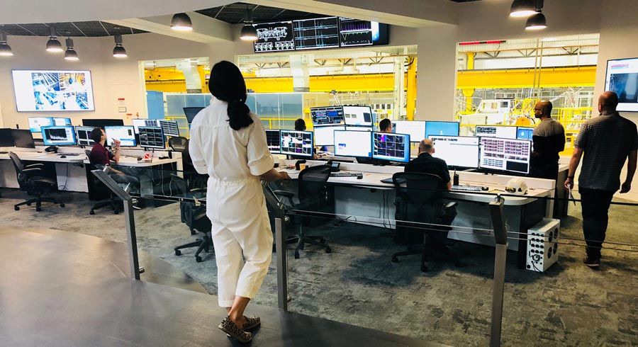 The control room at TAE. In the background, the reactor is visible though large windows. People are seated at desks with multiple monitors in the room, and a woman stands in the foreground.